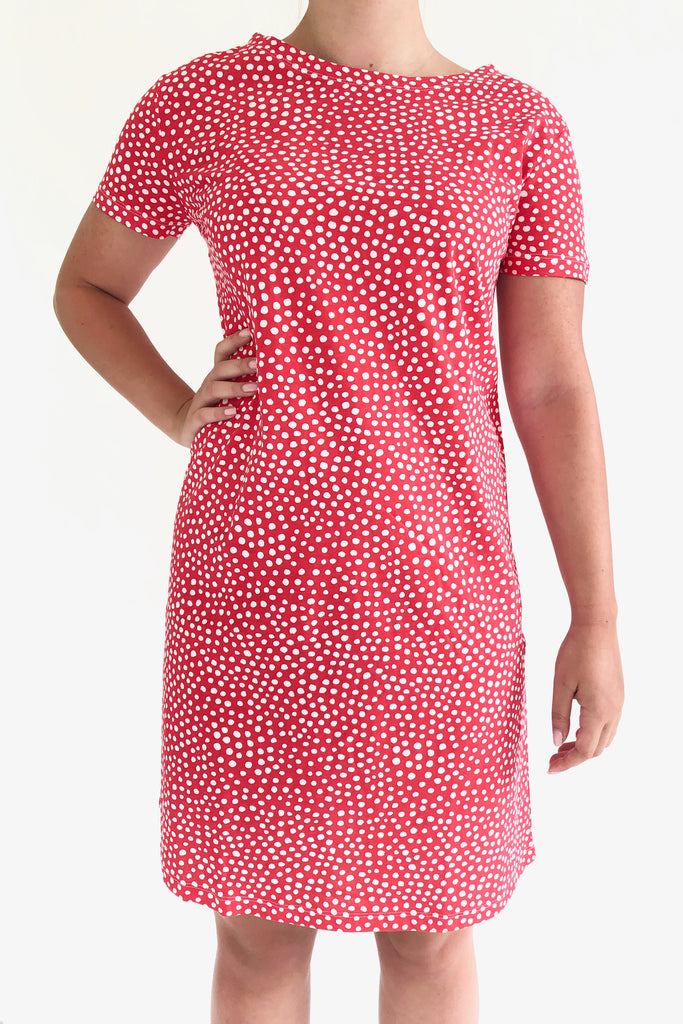 Person wearing a red dress with a white polka dot pattern, made from 100% cotton, standing with one hand on their hip against a plain white background. The dress is the Knit Dress Short Sleeve by See Design.