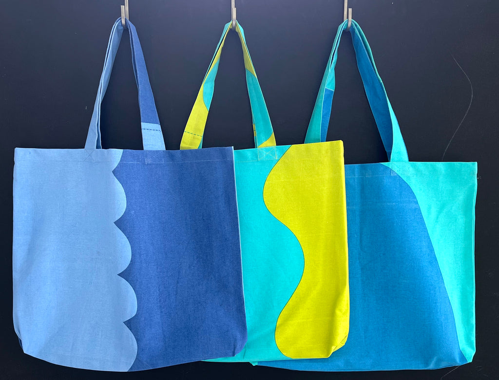 Three hand-painted blue and yellow cotton canvas Graphic Tote Bags hanging on a black wall.