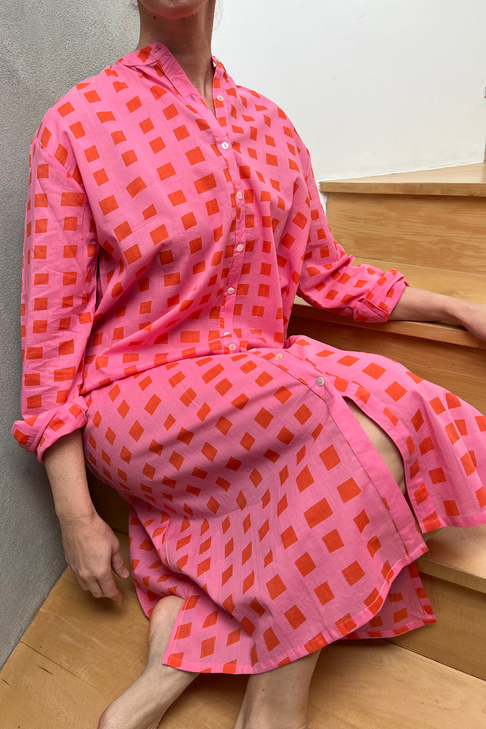 A woman in a pink checkered See Design shirt dress sitting on the stairs, showcasing both style and comfort.