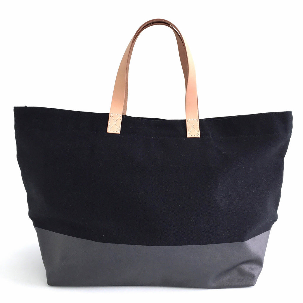 The Overnighter Bag by See Design is a fashionable two-toned tote with black upper and gray lower sections, adorned with light brown leather handles. Crafted from durable cotton canvas, this versatile bag is an ideal companion for carry-ons or overnight stays.