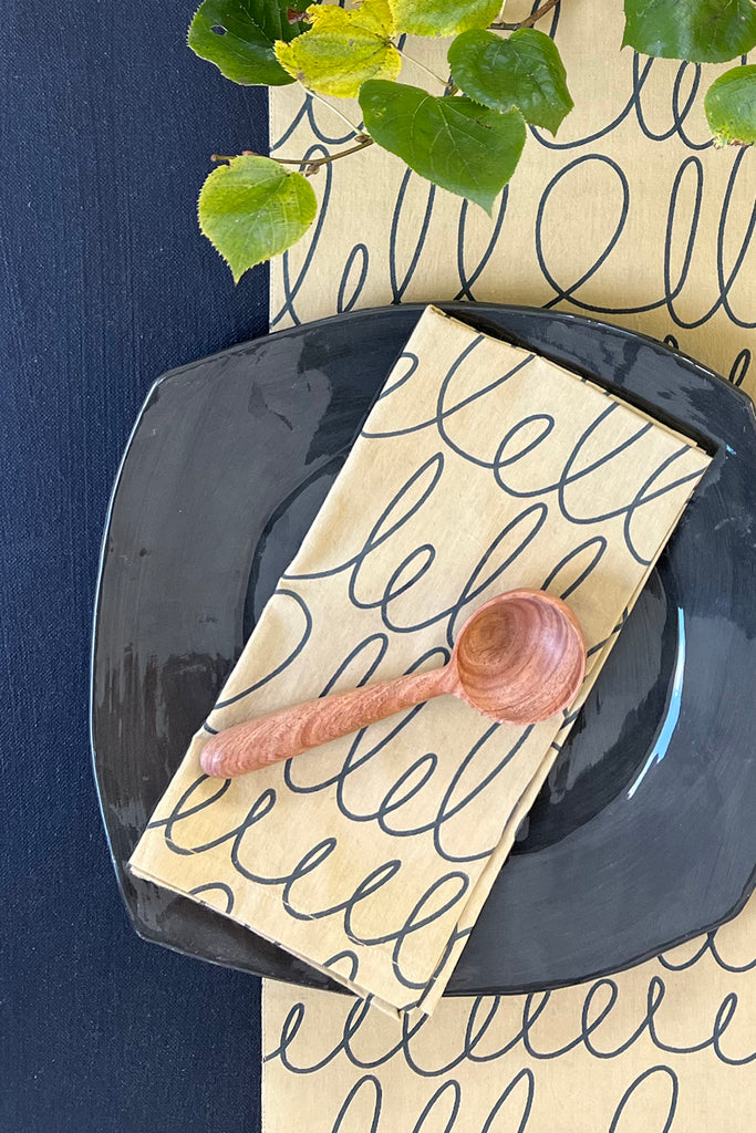 A See Design Napkins (Set of 4) in cotton with a looped pattern rests under a wooden spoon on a black plate and table runner, while green leaves in the top right enhance the fresh, elegant look.