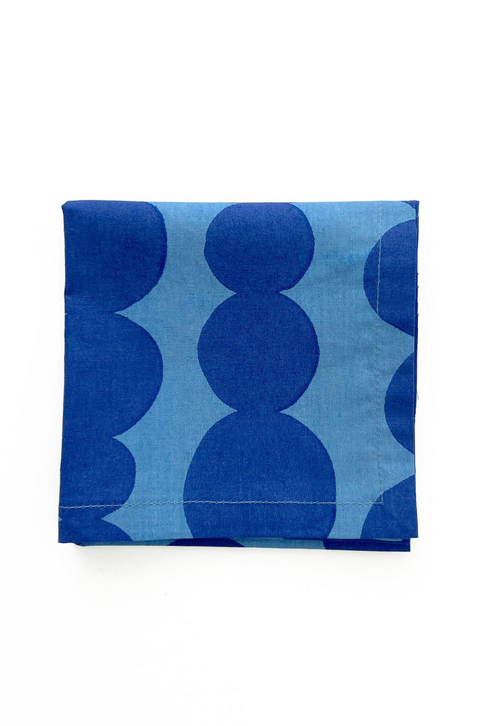 A set of four cocktail napkins by See Design, featuring a large circular pattern in blue, reminiscent of hand-painted designs, is folded neatly on a white background.