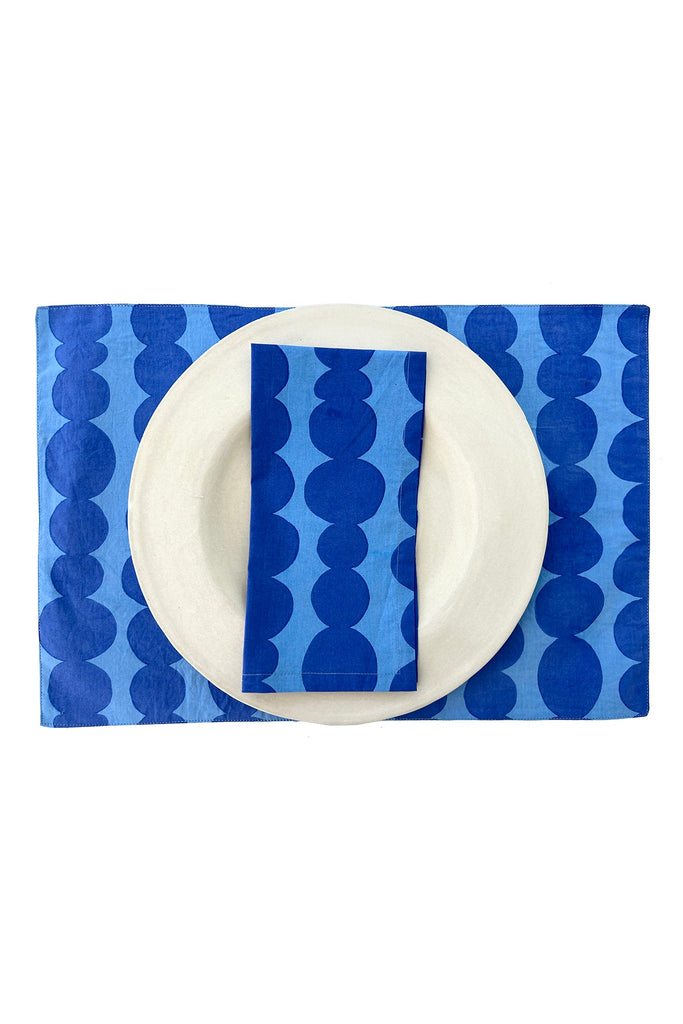 A white plate with a matching blue napkin on top, elegantly arranged on See Design's cotton placemats (set of 2) featuring a light blue and dark blue polka-dot pattern, enhances your dining experience.