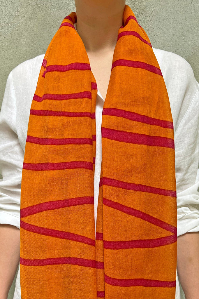 A person wearing a white shirt is shown adorned with the See Design Wool Scarf, featuring red horizontal and diagonal stripes on an orange background, bringing vibrant colors to their outfit.