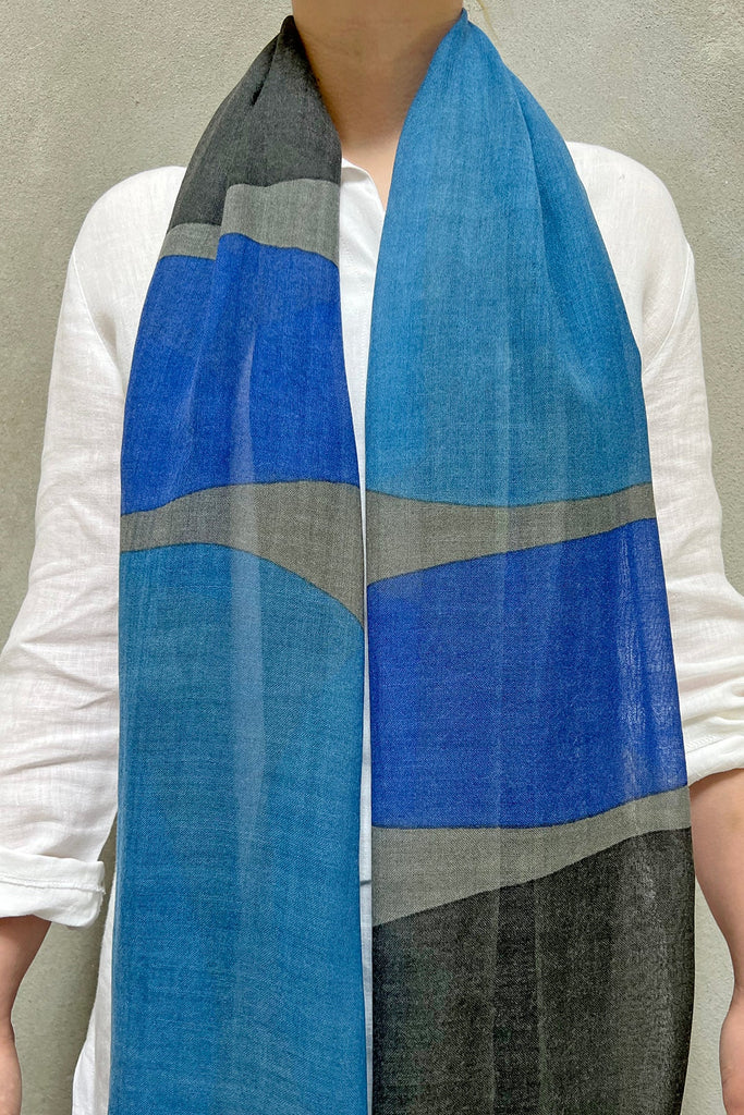 Person wearing a white shirt and a See Design wool scarf adorned with vibrant colors of blue, grey, and black stripes against a plain background.