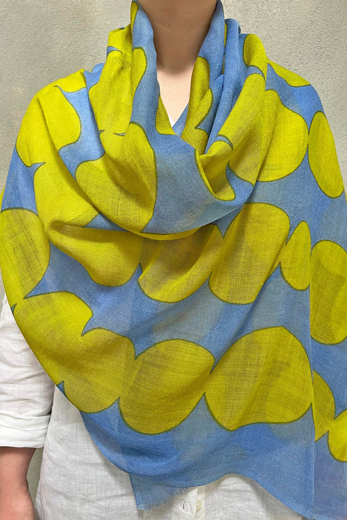 A person is wearing a white top and a See Design wool scarf, featuring a blue and yellow pattern, draped over their shoulders. The scarf, perfect for those seeking vibrant colors, showcases an abstract design with wavy, overlapping shapes. This wool scarf effortlessly adds flair to any outfit.