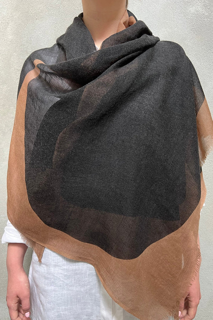 A person stands against a plain background wearing the See Design Wool Scarf in large, draped sections of dark gray and brown over a white garment.