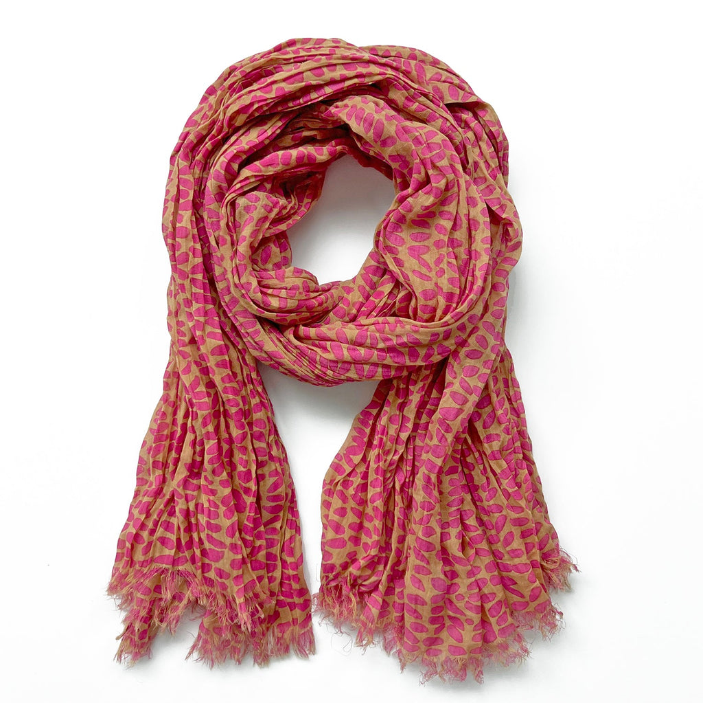 The Cotton Scarf by See Design is a soft, lightweight accessory in pink and beige, featuring a charming leaf pattern and fringed edges, skillfully folded into a circular shape.