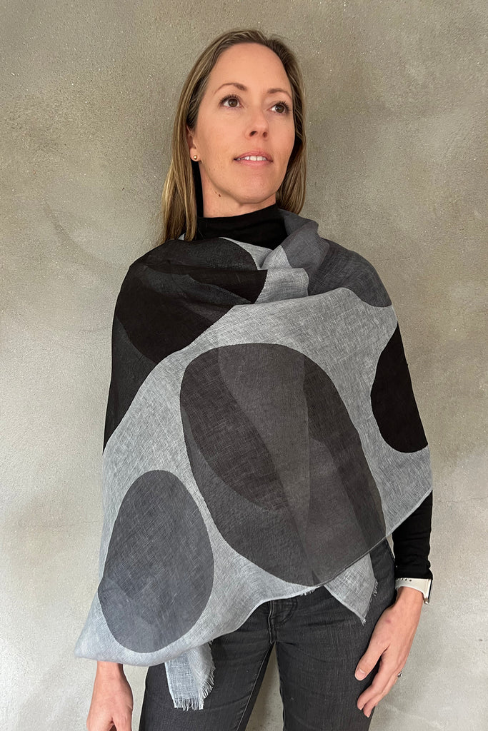 A woman poses against a neutral background, wearing a black top and the See Design Linen Scarf featuring gray and black abstract circular patterns.