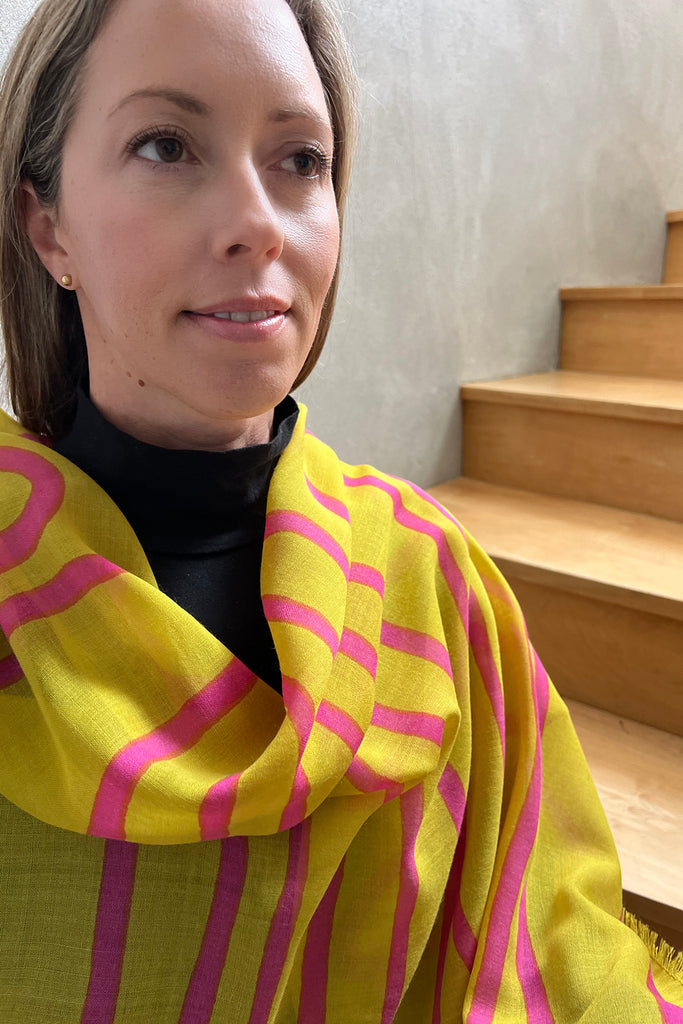 A woman wearing a yellow Wool Scarf with pink stripes from See Design and a black top sits beside a wooden staircase, gazing slightly to the side.