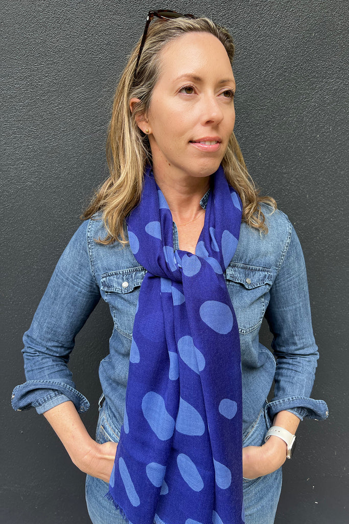 A woman wearing a See Design Wool Scarf and a denim shirt stands against a dark gray wall, looking slightly to the side with her hands in her pockets.