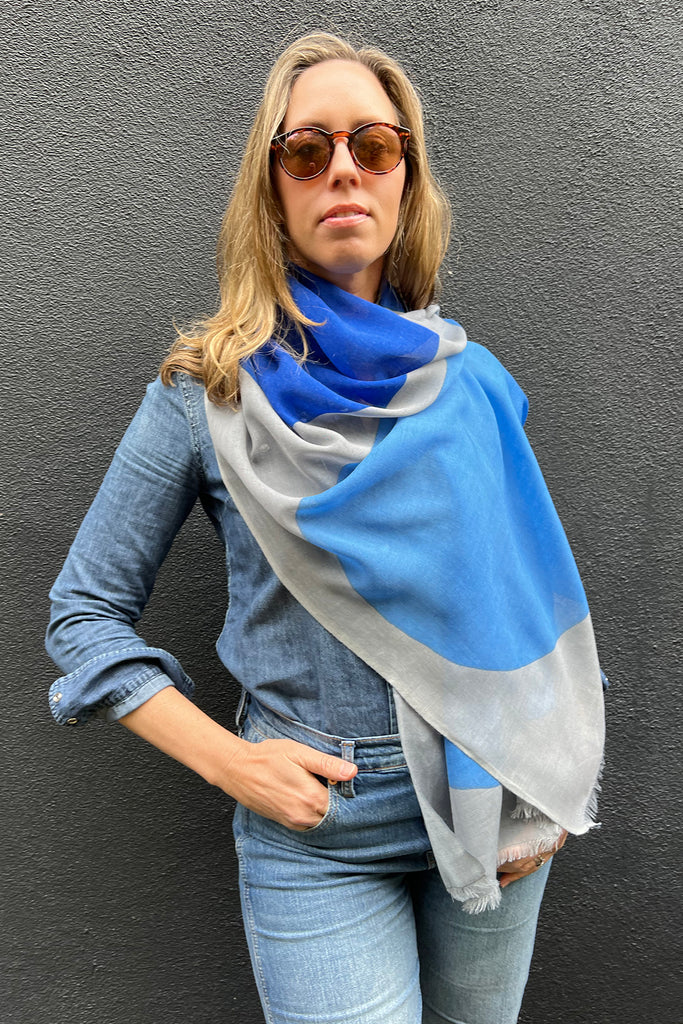 A woman in sunglasses, a denim shirt, and jeans stands against a dark wall, draped in a large blue and gray See Design Wool Scarf.