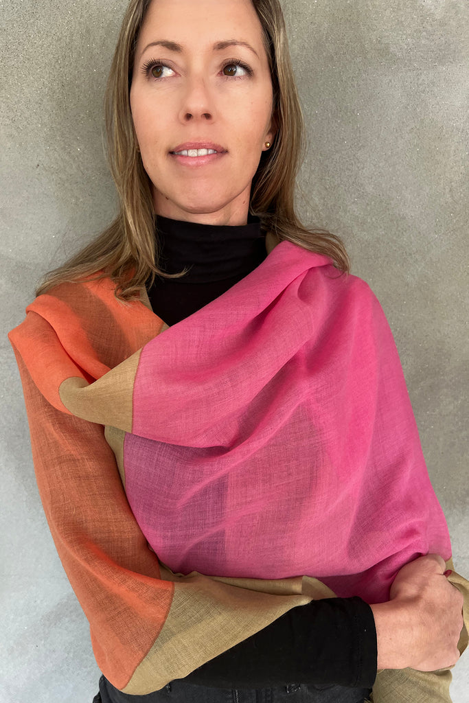 A woman with long hair, wearing a black top and a See Design Wool Scarf in pink, orange, and beige by Donna Gorman, stands against a gray background.