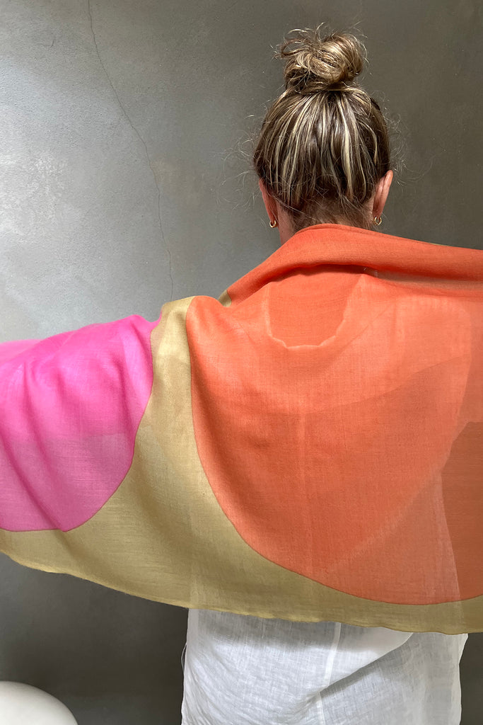A person with blonde hair in a bun, wearing a white top, drapes the See Design Wool Scarf—featuring orange, pink, and beige hues—over their shoulders while facing a gray wall.