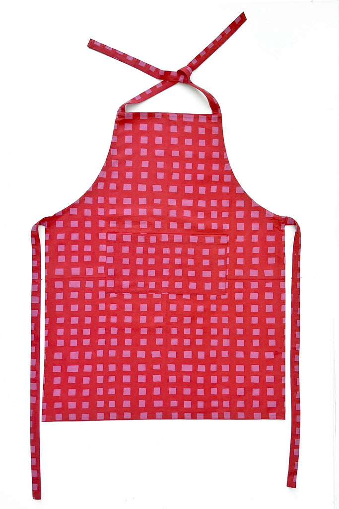 See Design Aprons: Red kitchen apron featuring hand-painted pink square patterns, a neck loop, and long waist ties, shown on a white background.