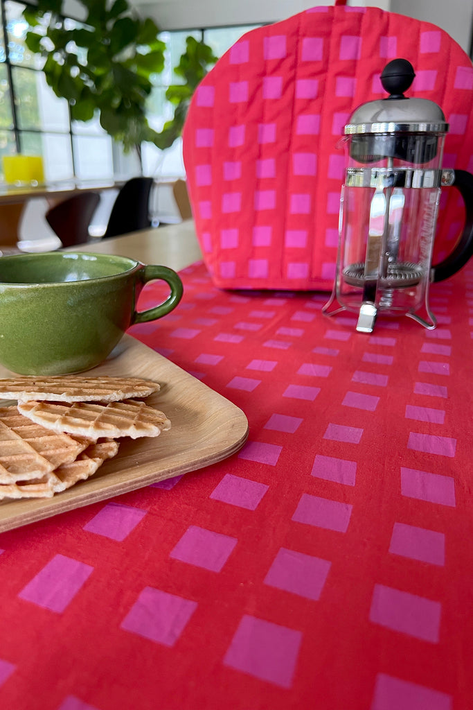 A green cup and saucer with wafer cookies on a wooden tray, a French press, and a See Design Tea Cosy accent the red patterned tablecloth—an inviting touch for your fall home collection.