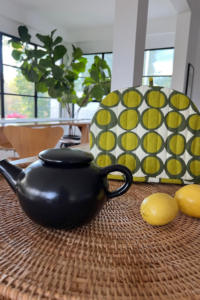 A black teapot, two lemons, and a green patterned Tea Cosy from See Design are arranged on a woven surface in a bright dining area with large windows and a leafy plant.