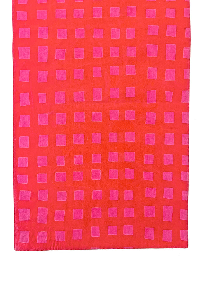 The See Design Table Runner features red cotton fabric with a repeating pattern of pink squares, making it perfect for adding a pop of color to your table setting.