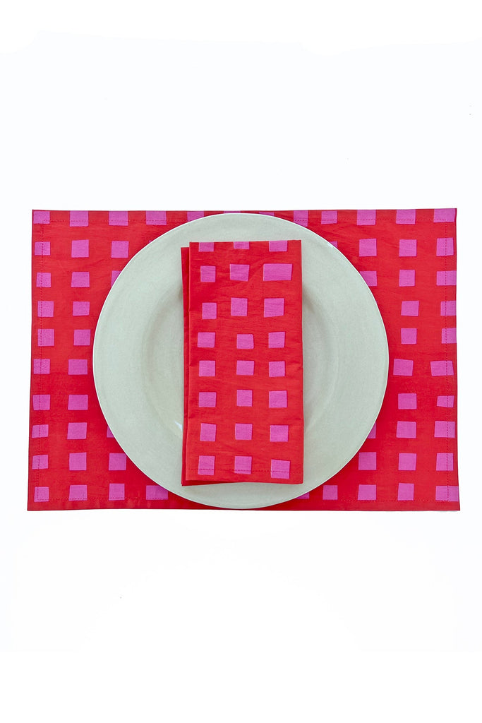 A white plate rests on See Design’s Placemats (Set of 2) in red and pink checks, with a matching napkin, all elegantly arranged on a white background.