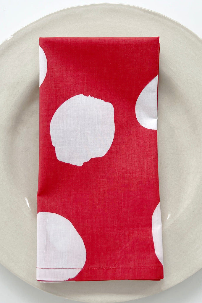 A set of 4 red cotton napkins with large white circles from See Design is neatly folded on a round off-white plate, bringing a stylish pop of color to your table.