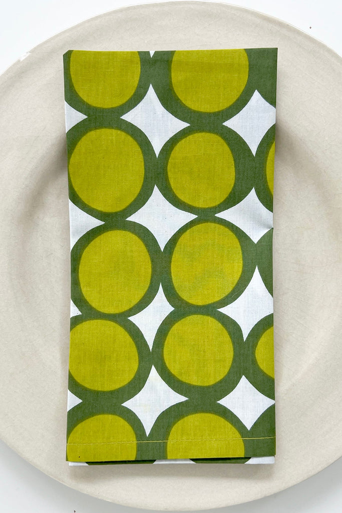 A white plate with a folded See Design Napkin (Set of 4) featuring a green and yellow geometric circle pattern, adding a vibrant accent to your colorful table linens.