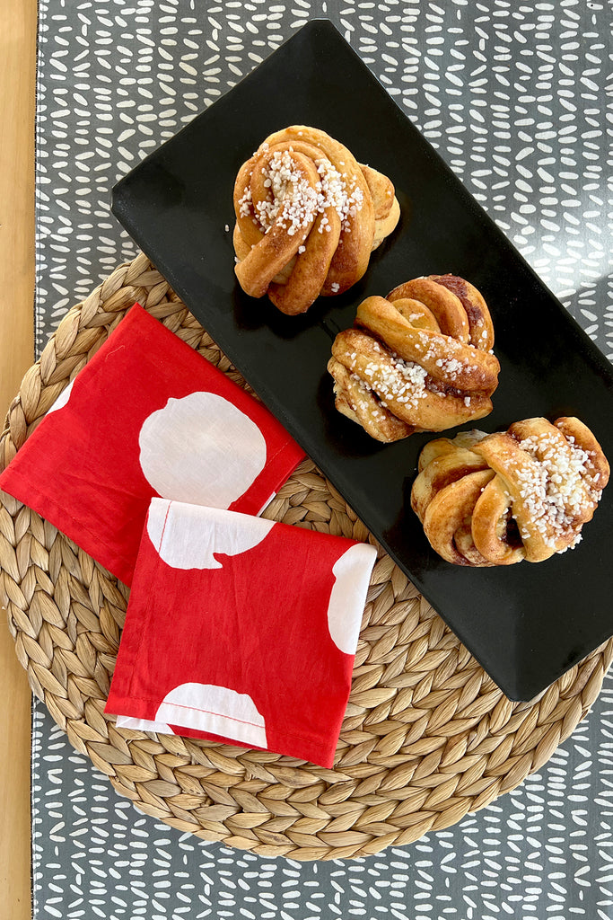 Three cinnamon buns with pearl sugar are set on a black rectangular plate, alongside See Design's red and white Cocktail Napkins (Set of 4), displayed on a woven placemat.