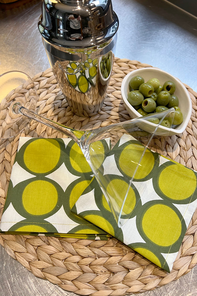 A martini glass, cocktail shaker, bowl of green olives, and a set of 4 See Design Cocktail Napkins with hand-painted patterns are arranged on a woven placemat.