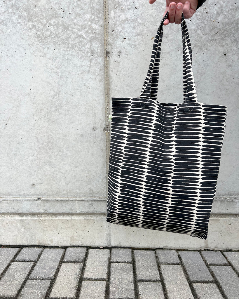 A hand holds an Easy Tote Bag from See Design, featuring black and white stripes on a lightweight cotton canvas, set against a concrete wall background with a paved surface below.