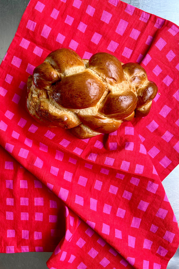A loaf of braided bread rests on a See Design Tea Towel (Set of 2) featuring a pink square pattern, by Donna Gorman, displayed on a metal surface.
