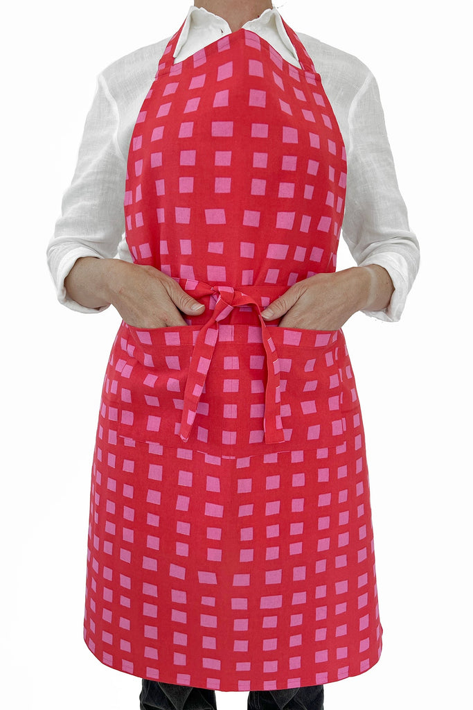 A person ties a See Design Aprons brand red and pink checkered apron over a white shirt at the waist.