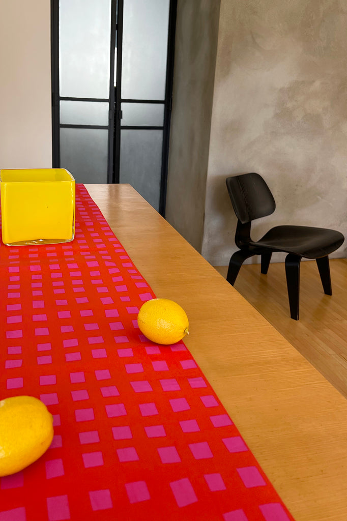 A wooden table with a See Design Table Runner in red and pink cotton, three lemons, a yellow square container, and a black chair creates a charming table setting in a modern room.