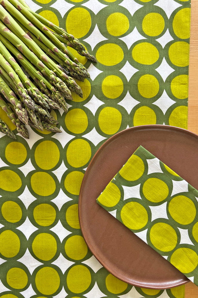 A bundle of asparagus, a brown plate with a matching napkin, and the See Design Table Runner create a fresh table setting with a green and yellow geometric circle pattern.