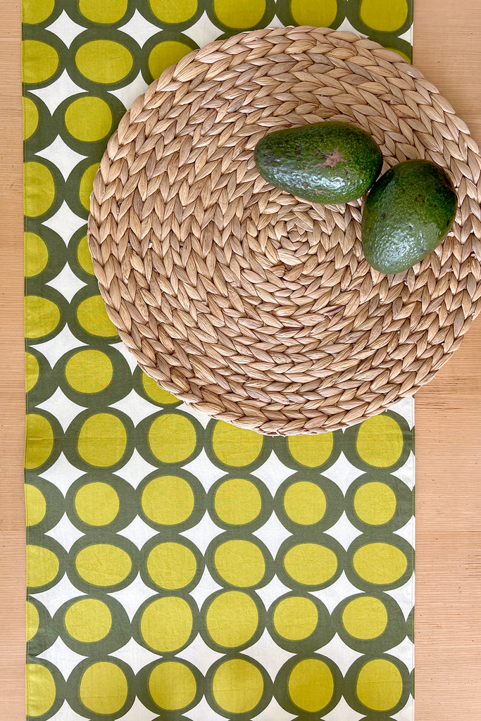 A woven circular placemat with two avocados sits on a See Design Table Runner, which features a vibrant green and yellow geometric circle pattern.