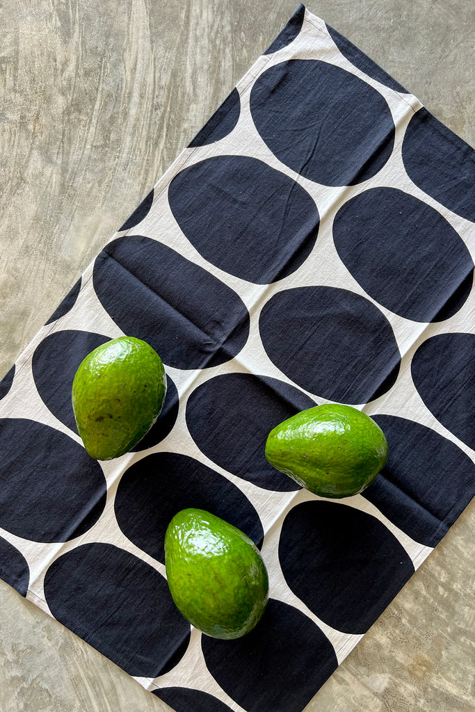 Three green avocados rest on a See Design tea towel from the Tea Towels (Set of 2), its black-and-white circular patterns adding charm to the kitchen setting.
