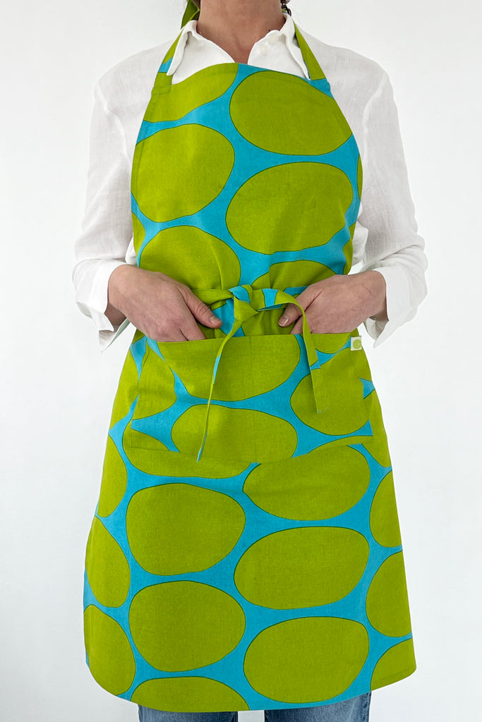 A person in a white shirt and blue jeans is tying an apron by See Design, featuring vibrant blue oval patterns.