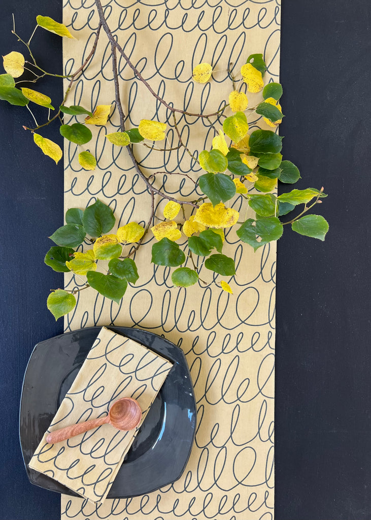 A black plate with a wooden spoon rests on a napkin and See Design Table Runner featuring a looped pattern; above, a branch with green and yellow leaves adds a natural touch to the setting.
