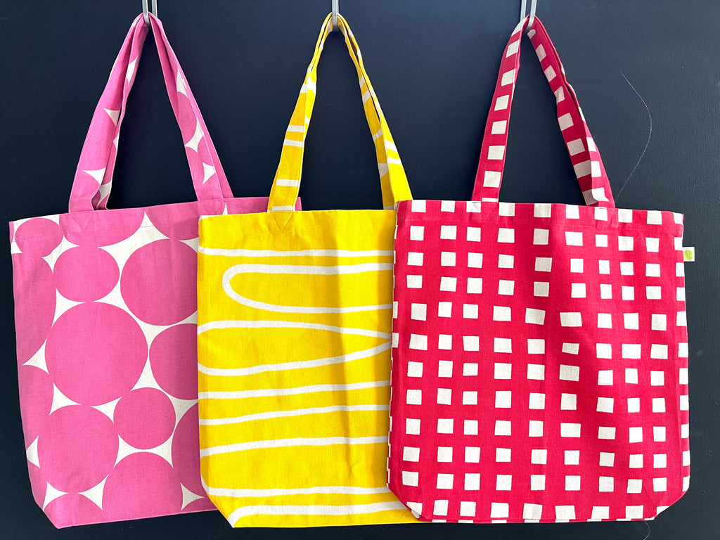 Three lightweight, hand-painted cotton canvas Easy Tote Bags hanging on a black wall by See Design.