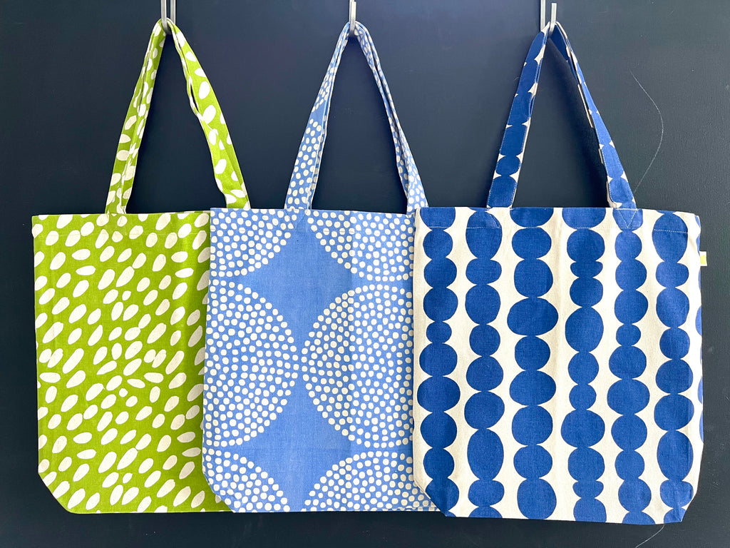 Three Easy Tote Bags with hand-painted artwork hanging on a black wall.