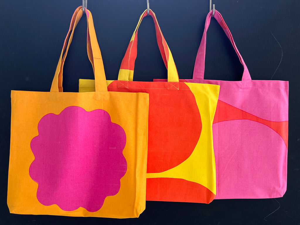 Three See Design Graphic Tote Bags in vivid hues hanging on a black wall.