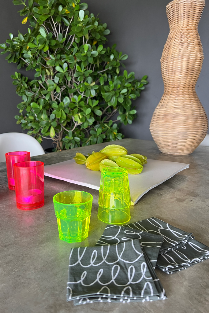 Colorful plastic cups sit on a table with hand-painted designs, while a plant is next to a wicker vase. A set of See Design cocktail napkins shares space with star fruits on a tray.