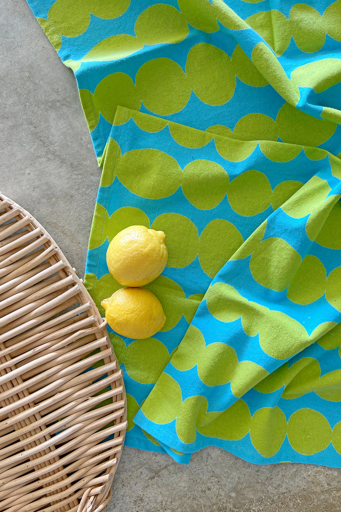 A vibrant blue and green polka dot tea towel (set of 2) made of cotton, next to a basket of fresh lemons. Brand name: See Design.