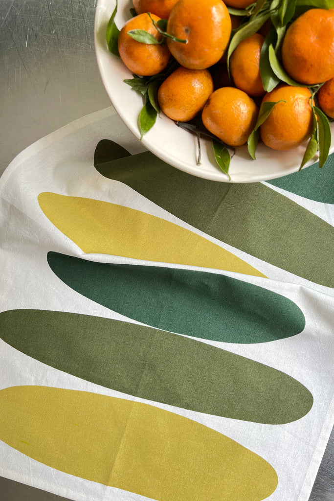 A bowl of vibrant oranges on a table next to a See Design Tea Towels (Set of 2).