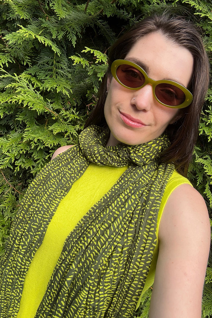 A person wearing green sunglasses, a yellow top, and a See Design Cotton Scarf stands in front of green foliage.