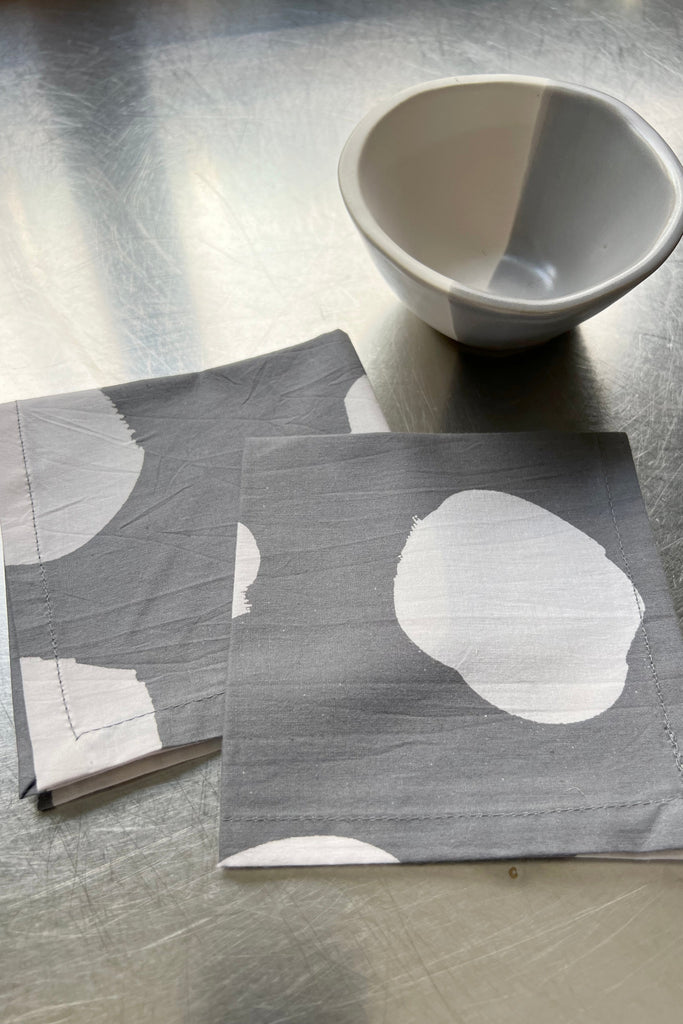 Two cocktail napkins from the See Design set of four, adorned with white circles, are placed on a metal surface next to a small bowl that is white and gray.
