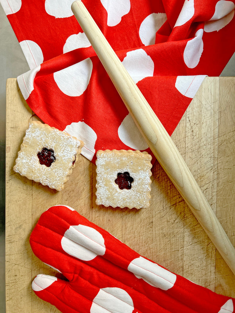 A wooden rolling pin, two square jam-filled cookies dusted with powdered sugar, a red polka dot oven mitt, and See Design Tea Towels (Set of 2) are displayed on a wooden surface.