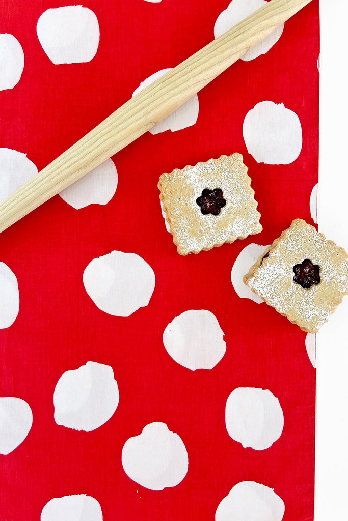 A wooden rolling pin and two powdered sugar-dusted sandwich cookies sit atop a See Design Table Runner, featuring red cotton with white polka dots for a charming table setting.