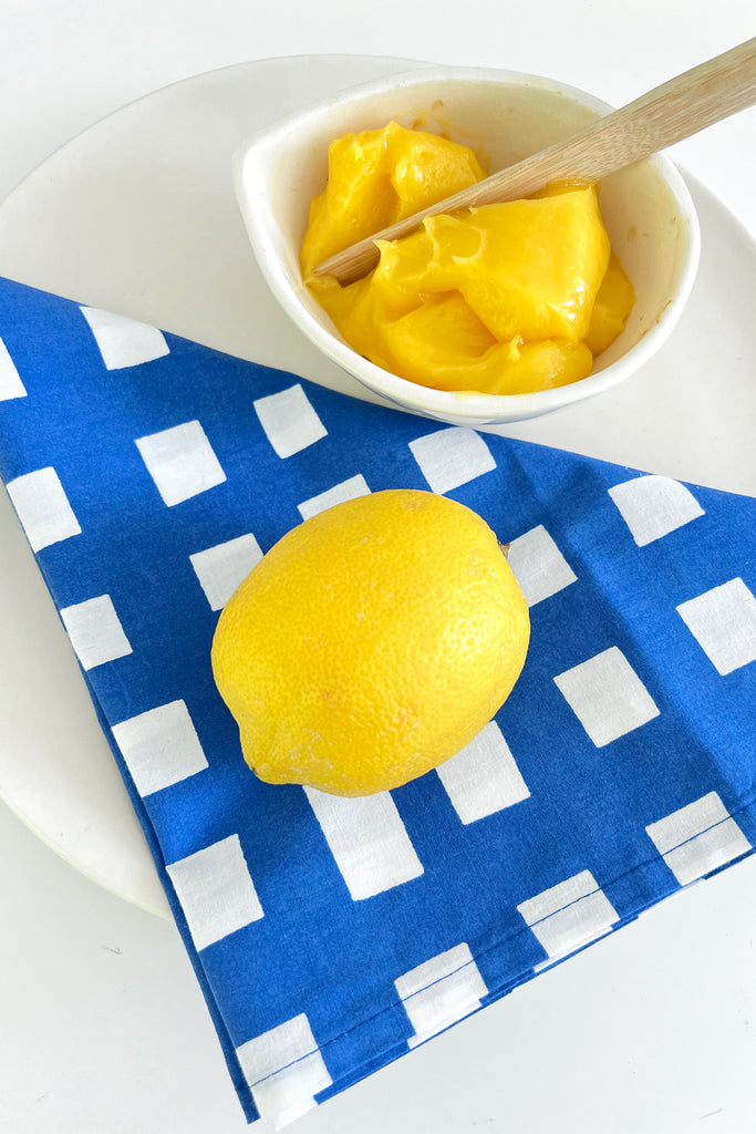 A lemon rests on a blue and white checkered See Design Napkins (Set of 4). Next to it, a small bowl filled with lemon curd has a wooden spoon in it. The items are placed on a white surface.