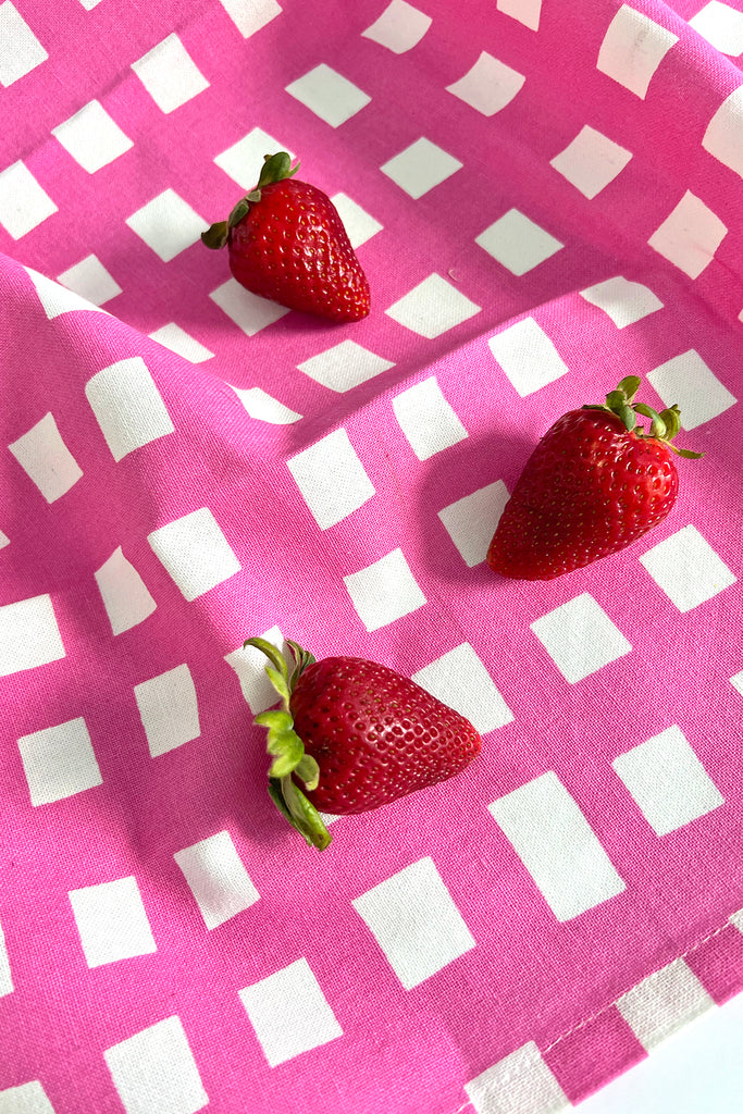 Three strawberries rest on a pink and white checkered cloth, reminiscent of See Design's Tea Towels (Set of 2) in a charming kitchen setting.