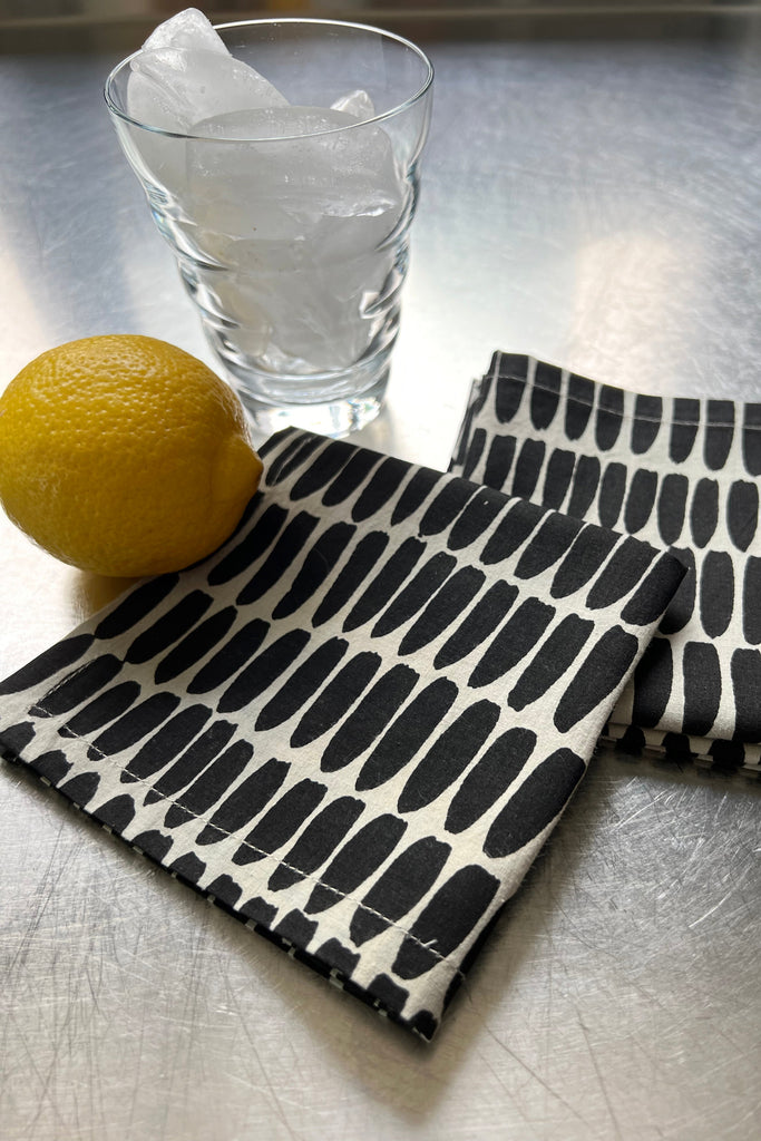 A glass filled with ice, a lemon, and two of See Design's black and white patterned Cocktail Napkins (Set of 4) are placed on a metallic surface.