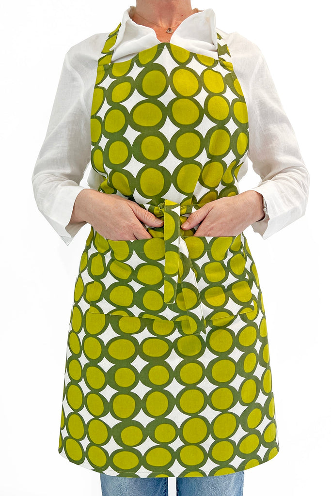 A person wearing a white shirt and blue jeans ties a See Design green and yellow hand-painted patterned kitchen apron around their waist, standing against a plain white background.